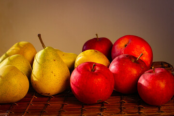Several ripe yellow pears and red apples stand on a wicker basket illuminated by rays of the sun