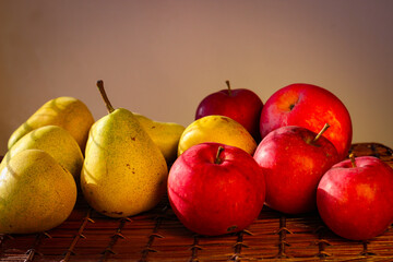 Several ripe yellow pears and red apples stand on a wicker basket illuminated by rays of the sun