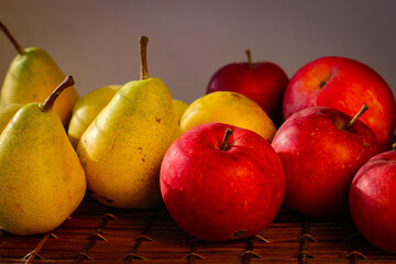 Several ripe yellow pears and red apples stand on a wicker basket illuminated by rays of the sun