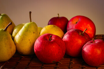 Several ripe yellow pears and red apples stand on a wicker basket illuminated by rays of the sun
