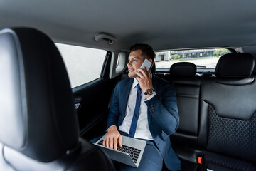 Smiling businessman talking on smartphone and using laptop in car.