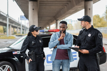 police officers looking at african american victim thinking near patrol car on blurred background...