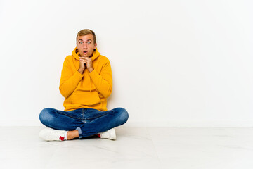 Young caucasian man sitting on the floor praying for luck, amazed and opening mouth looking to front.