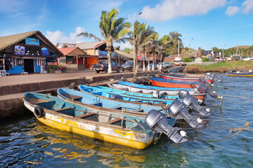 Colorful boats in the port of Hanga Roa, near Plaza Hotu Matua, surrounded by palm trees, against a blue sky.