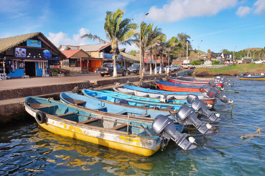 Colorful boats in the port of Hanga Roa, near Plaza Hotu Matua, surrounded by palm trees, against a blue sky.