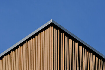 Fototapeta premium Detail of the roof of a modern building with vertical larch slats cladding against bright blue sky. Perspective view from below.