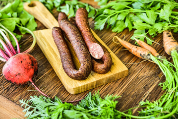 Sausage served on an oak board among vegetables from the home garden. - obrazy, fototapety, plakaty