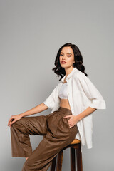 Curly woman with hand in pocket looking at camera while posing on stool isolated on grey