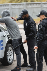 african american policeman with truncheon frisking hooded offender near patrol car with blurred colleague on foreground outdoors.