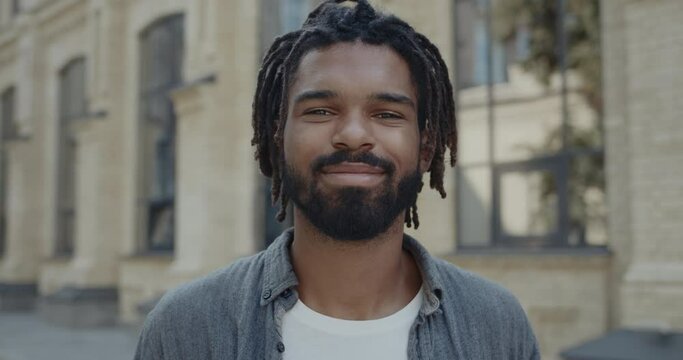 Close up of cheerful guy with beard turning head and looking to camera. Portrait of man with dreadlocks wearing casual clothes smiling while standing at city street. Concept of lifestyle