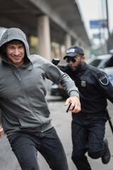 Fototapeta premium hooded offender running from african american police officer aiming with gun on blurred background outdoors.