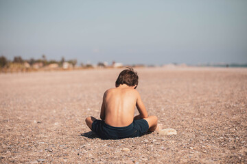Fototapeta na wymiar A boy without a shirt sitting on his back plays alone sitting on his back on the beach 