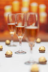 Three glasses of champagne on a white table, the second is in focus. The background is out of focus, one can sense that it is a fireplace with burning candles. There are chocolates on the table.
