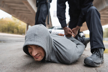 african american policeman handcuffing offender lying on street on blurred background outdoors.