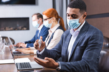 African american businessman in medical mask applying sanitizer on hands at workplace on blurred background.