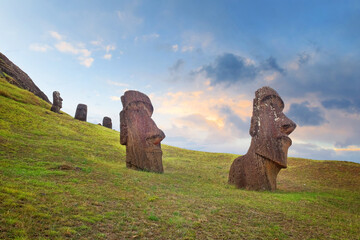 Moai on the slopes of the Rano Raraku Volcano, on Easer Island, surrounded by green vegetation, against a colorful sky.