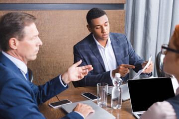 African american businessman pointing with finger at paper while sitting at workplace near blurred colleagues on foreground.