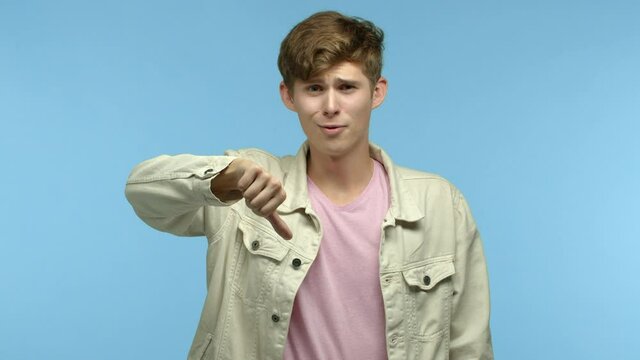 Arrogant guy mocking someone, showing thumbs down gesture and shaking head with disappointment, standing skeptical over blue background