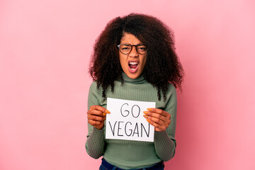 Young african american curly woman holding a go vegan placard screaming very angry and aggressive.