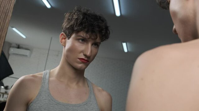 Confused and angry trans man stares in the mirror. Androgynous guy with makeup looks at his reflection. Gender identity and social label concept 4k