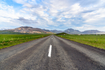 Empty road through a grassy valley in Iceland on a partly cloudy summer evening.