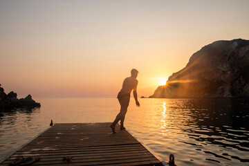 Fototapeta premium Silhouette of a man turned from behind while diving from a small marina on the sea. breathtaking sunset, golden hour.
