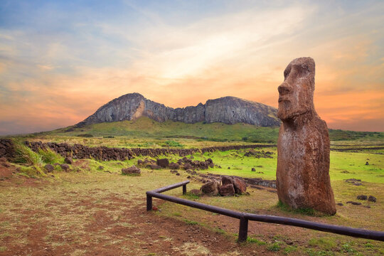 Stone fence and Moai statues at the entrance in the Ahu Tongariki ceremonial center on Easter Island, covered by a colorful sunset sky, against the crater of the Rano Raraku volcano.