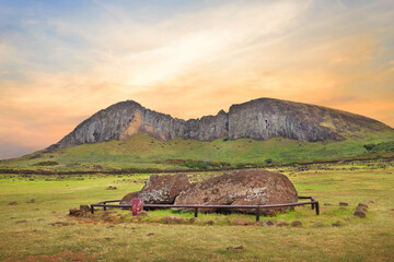Fallen Moai statue at the Ahu Tongariki ceremonial center on Easter Island, covered by a colorful sunset sky, against the crater of the Rano Raraku volcano.