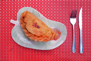 Chilean fried turnover called Empanada, consisting of pastry and tuna filling, served with a spicy sauce, traditionally eaten on Easter Island, on a red table cover made of plastic.
