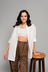  brunette woman in white top and shirt looking at camera while leaning on stool isolated on grey