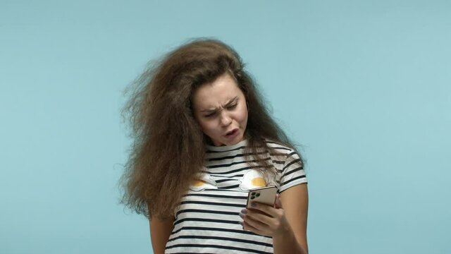 Funky girl with thick long hair reading smartphone with puzzled face, stare confused at camera and asking what, cant understand what happened, blue background