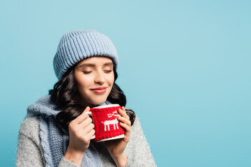  woman in hat and scarf, with closed eyes holding cup with knitted holder isolated on blue
