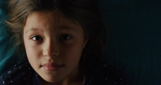 Cinematic shot of little girl is lying on bed and looking in camera while trying to fall asleep with soft atmospheric light at home at night.Concept of children,healthcare, insomnia, bedding, comfort.