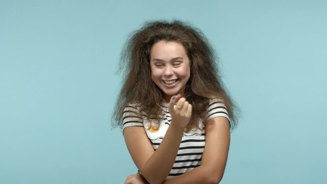 Young seductive woman beckon you flirty with finger, asking to come closer and smiling, luring someone, standing over blue background in striped t-shirt