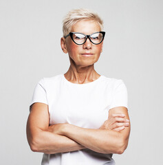 Old senior woman wear eyeglasses with her arms crossed over grey background.