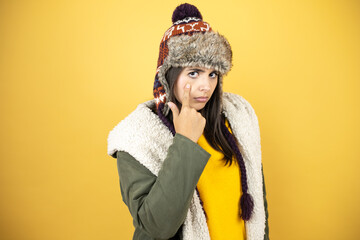 Young beautiful woman wearing a hat and a green winter coat over yellow background Pointing to the eye watching you gesture, suspicious expression