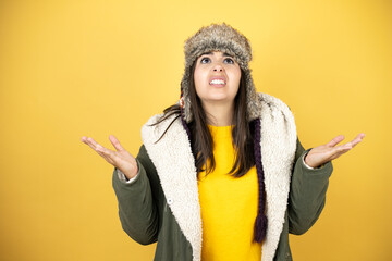 Young beautiful woman wearing a hat and a green winter coat over yellow background clueless and...