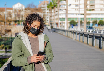 Young Attractive Curly Long Hair Boy With Black Mask and Green Jacket Writing Text Message on his Phone in a Bridge at Sunny Day