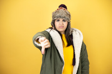 Young beautiful woman wearing a hat and a green winter coat over yellow background with angry face,...