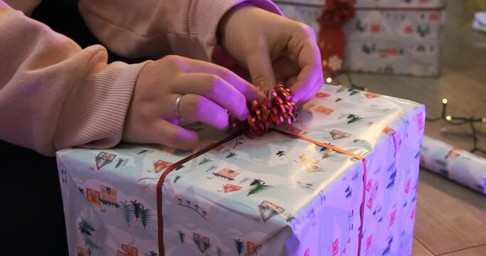 Girl female hand decoration a holiday Christmas gift box with a beautiful funny wrapper and thin red ribbon, tape with a bow in center. Decor paper and light on the background, family happy atmosphere