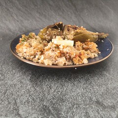 A blue plate with  an artichoke cutted in half with a quinoa salad side and a slice of lemon in a dark silver background