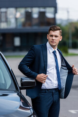 young businessman taking off blazer near car on blurred foreground.