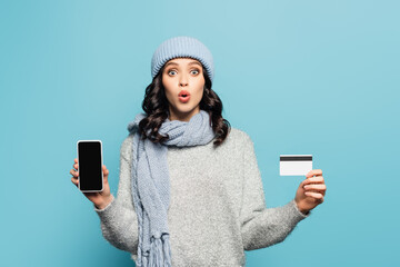Shocked woman in hat and scarf looking at camera while holding smartphone and credit card isolated on blue
