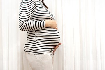  Closeup of young beautiful pregnant woman in striped t-shirt, that holding her belly and standing near window at home. Concept of pregnancy preparation. Mother with big belly, hands over tummy.