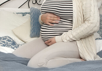 Closeup of young beautiful pregnant woman holding her belly and sitting on the bed. Concept of pregnancy preparation and expectation, gynecology. Mother with big belly, hands over tummy.