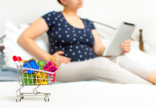 Closeup of shopping cart with colorful gift boxes on the front and pregnant woman with big tummy using a tablet on the background. Mother buy baby products. Shopping online during quarantine. - Powered by Adobe