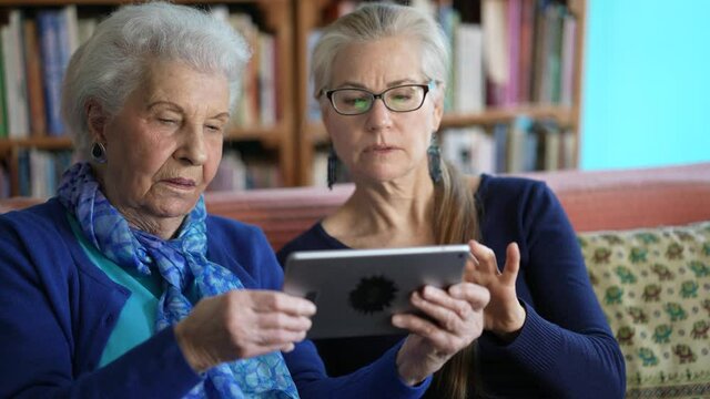 Happy Elderly Senior Woman And Mature Woman Look At Tablet Computer While Drinking Tea On A Couch In A Living Room.