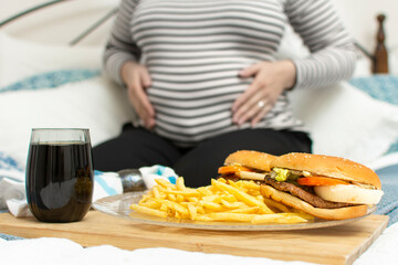 Cropped image of young hungry pregnant woman eating a hamburger and fried potatoes. Concept of unhealthy nutrition during pregnancy, Mother waiting baby. Close-up of belly and junk food (fast food). 