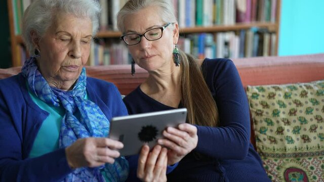 Happy elderly senior woman and mature woman look at tablet computer while drinking tea on a couch in a living room.
