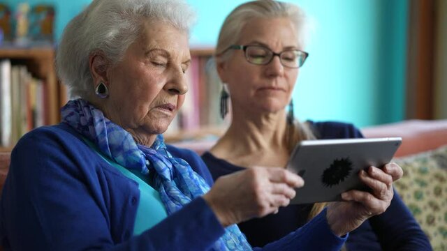 Happy elderly senior woman and mature woman look at tablet computer while drinking tea on a couch in a living room.